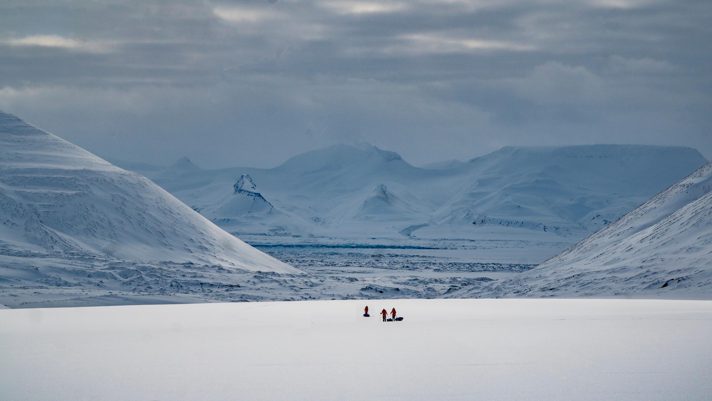Snow-covered mountains surround a vast white plain where three small figures traverse the icy landscape on sleds or skis.