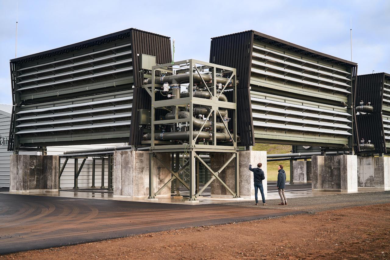 Modern industrial building with angular black structures and exposed metal framework. Two people stand nearby for scale.